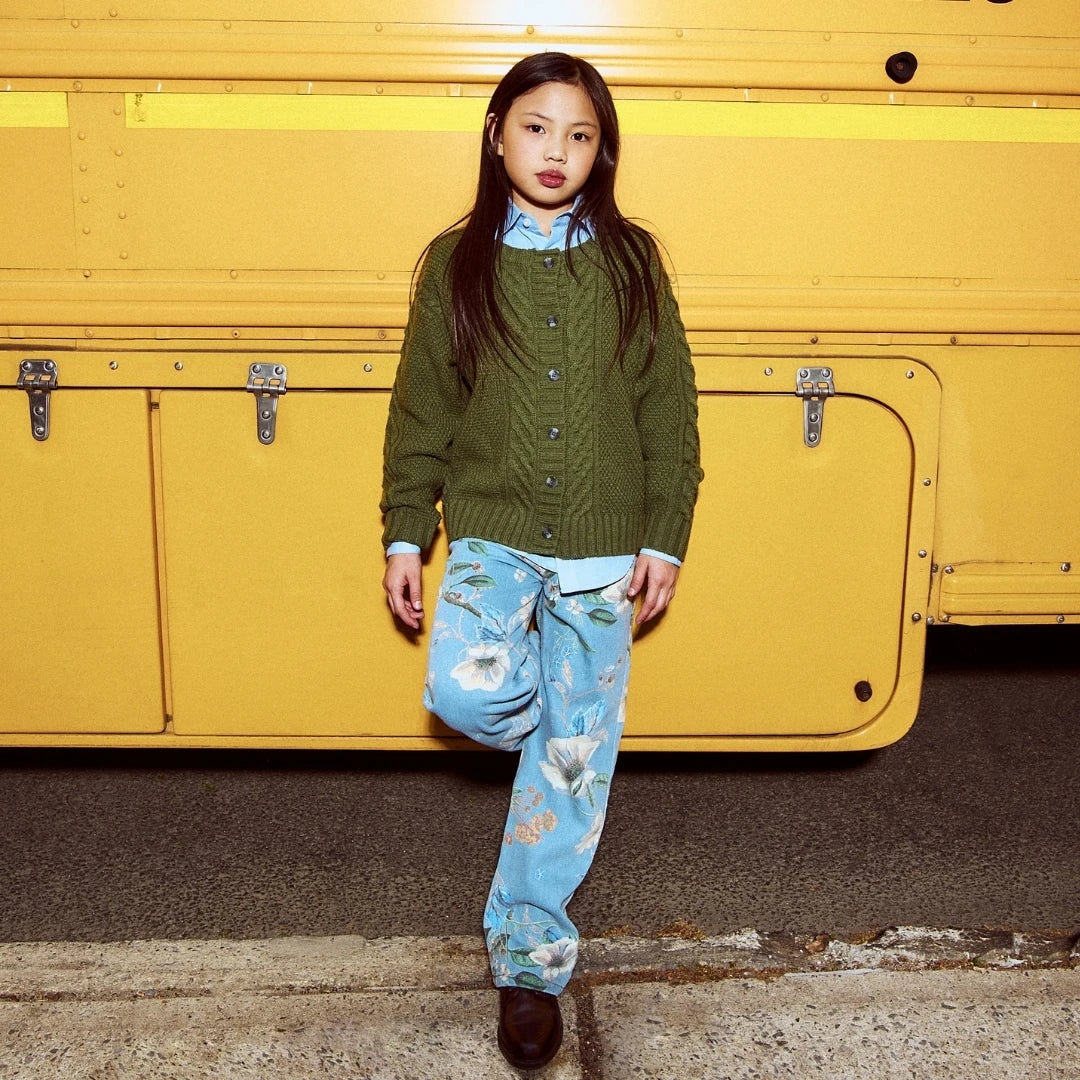 A young girl stands in front of a bright yellow bus, wearing an olive green cable-knit button-up sweater over a light blue collared shirt, paired with light blue floral pants and dark brown boots.