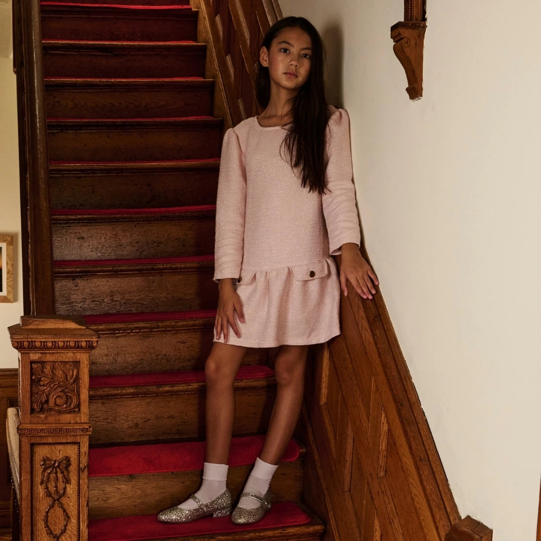 A young girl stands on a wooden staircase with red carpeting, wearing a light pink long-sleeve mini dress with a peplum hem, white socks, and sparkly silver flats.