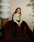 A girl wearing a cream sweater, seated on a velvet chair. Appaman Fall/Winter 2026 collection.