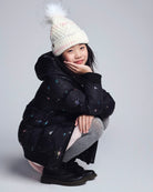 Child wearing a black puffer coat, gray pants, and a white knit hat with a pom-pom on a light gray background.
