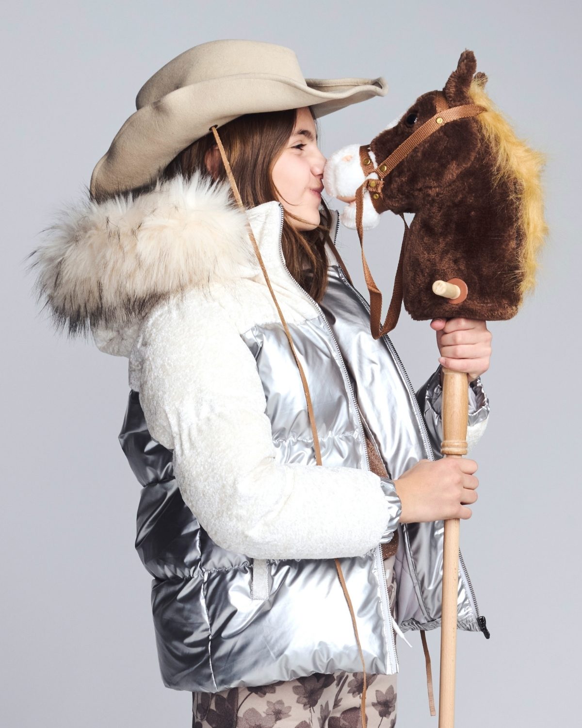 Person wearing a silver puffer jacket with fur hood, holding a plush horse toy against a gray background