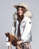 Person wearing a silver puffer jacket with fur hood holding a brown horse plush toy on a gray background