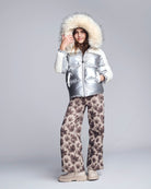 Person wearing a silver puffer jacket with a fur-lined hood and patterned pants on a gray background