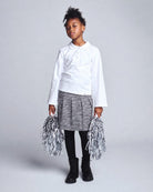 Child wearing a white shirt and gray skirt with pom-poms on a plain background