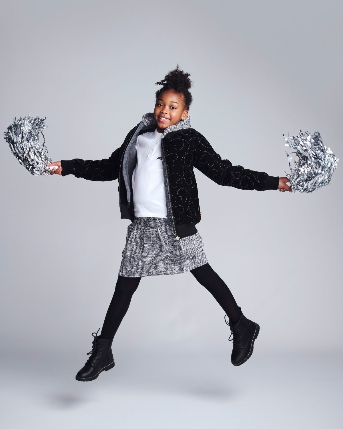 Child in black jacket and gray skirt holding silver pom-poms against a gray background
