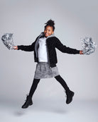 Child in black jacket and gray skirt holding silver pom-poms against a gray background