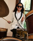 A young girl wearing black sunglasses, a white blazer with black lapels, and a white pleated shirt stands behind a drum set, holding drumsticks in an indoor room with a window.