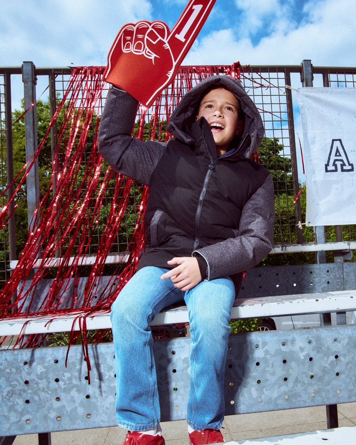 Child sitting on bleachers holding a red foam finger with a number one, outdoors.