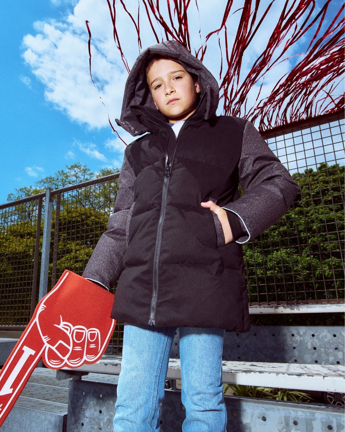 Child wearing a black puffer jacket standing outdoors with a red sign and tree branches in the background.