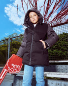 Child wearing a black puffer jacket standing outdoors with a red sign and tree branches in the background.