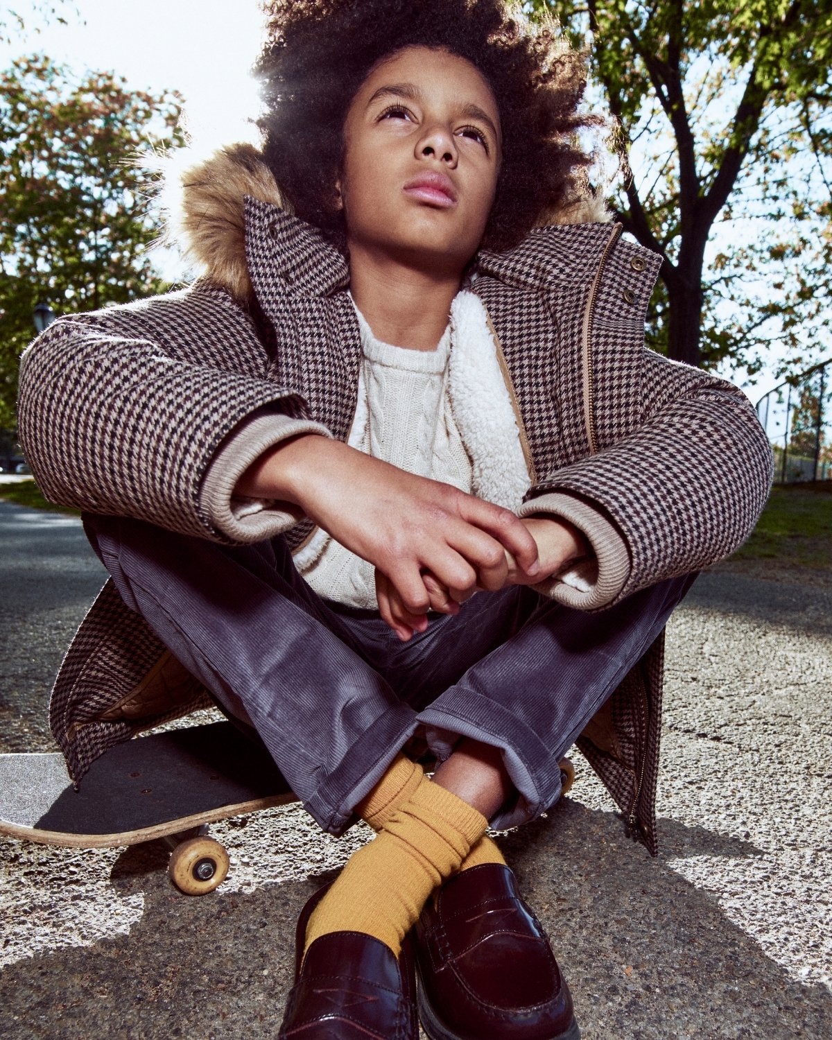 Child sitting on a skateboard wearing a plaid coat with fur hood, white sweater, and yellow socks.