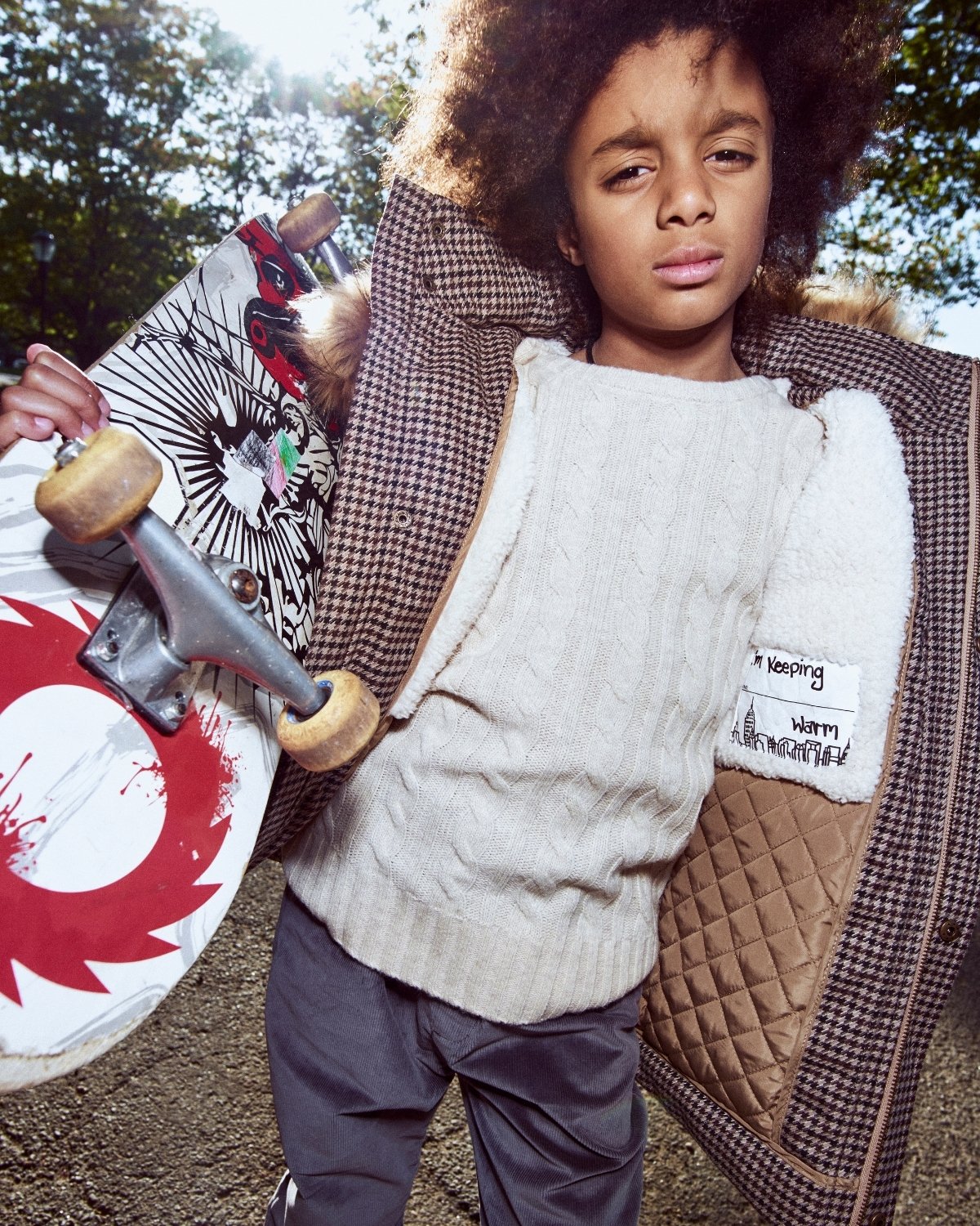 Child holding a skateboard outdoors with a checkered coat and patterned sweater.
