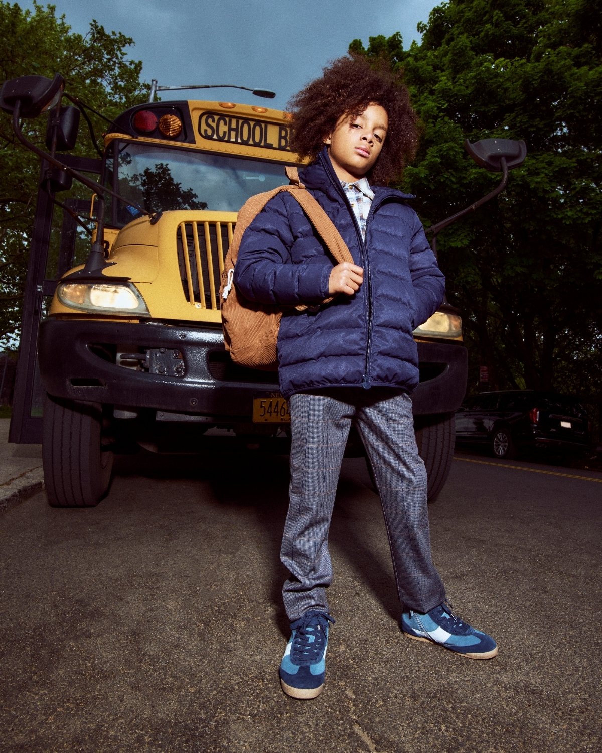 Child standing in front of a school bus wearing a blue jacket and gray pants.