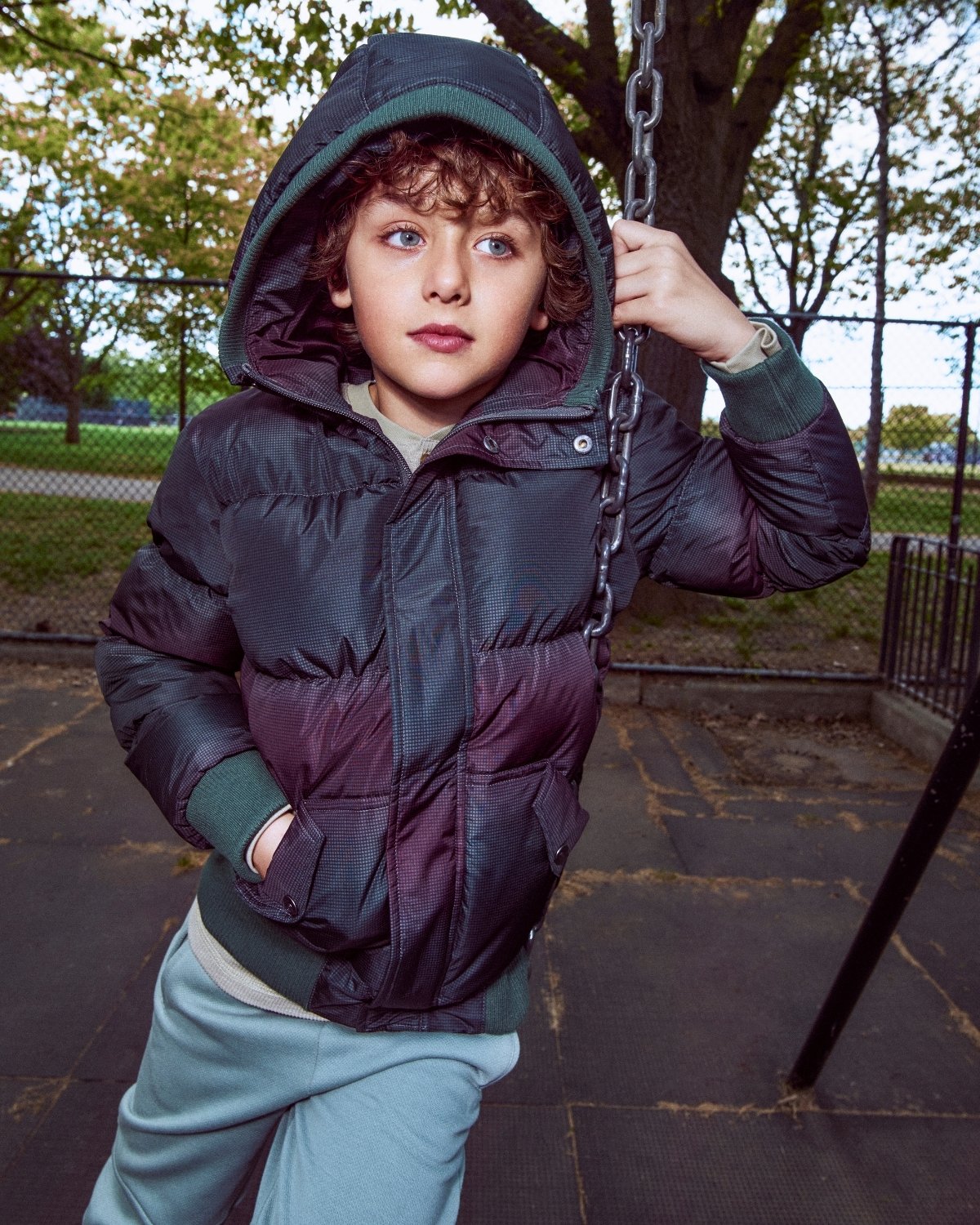 Child in a dark puffer jacket on a swing set with trees and playground equipment in the background.