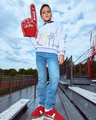 Child holding a foam finger with number 1, wearing a white sweatshirt and blue jeans, standing on bleachers.