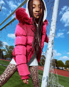Child in a pink puffer jacket and leopard print leggings standing near a soccer goal.