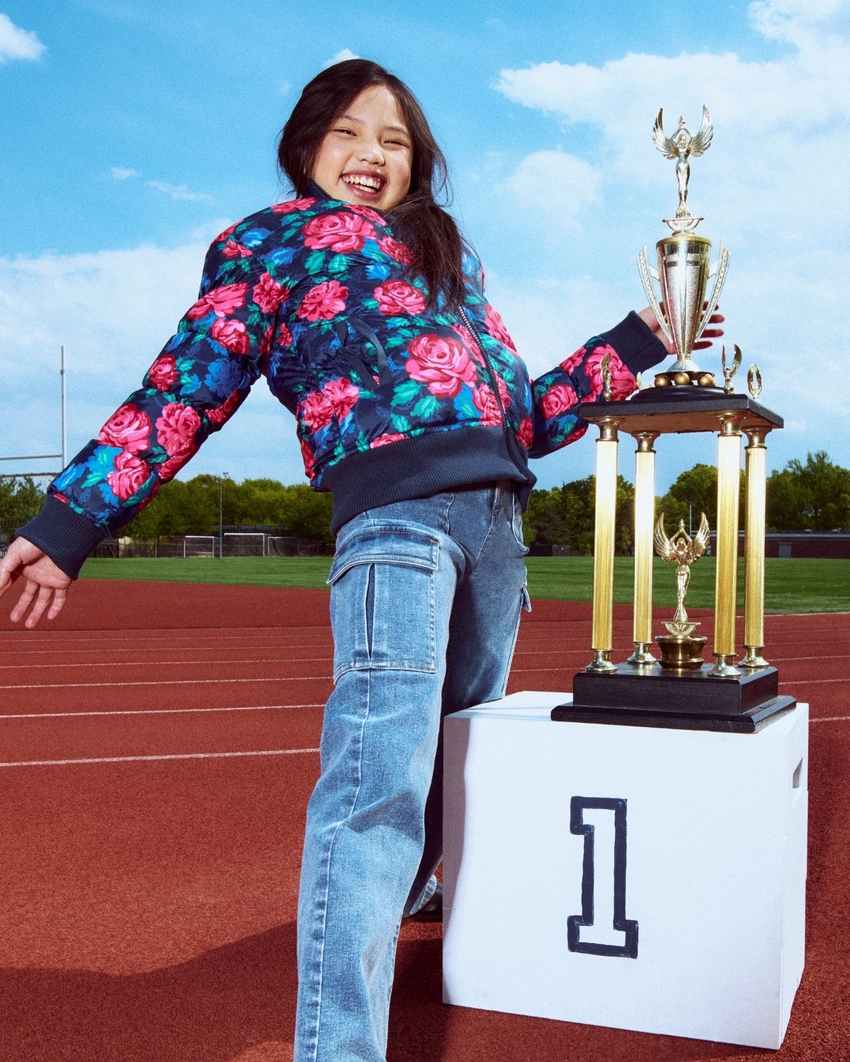 Person in floral jacket and jeans standing next to a trophy on a track field