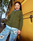 Woman in green cardigan and floral pants leaning against a yellow school bus.
