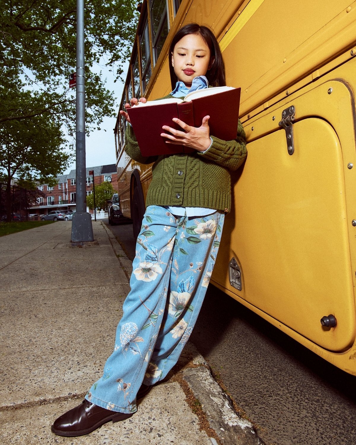 Young girl reading a book next to a yellow school bus on a sidewalk.