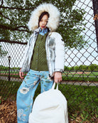 Person wearing a silver puffer jacket with a fur-lined hood, green sweater, and blue floral pants, holding a white bag outdoors.
