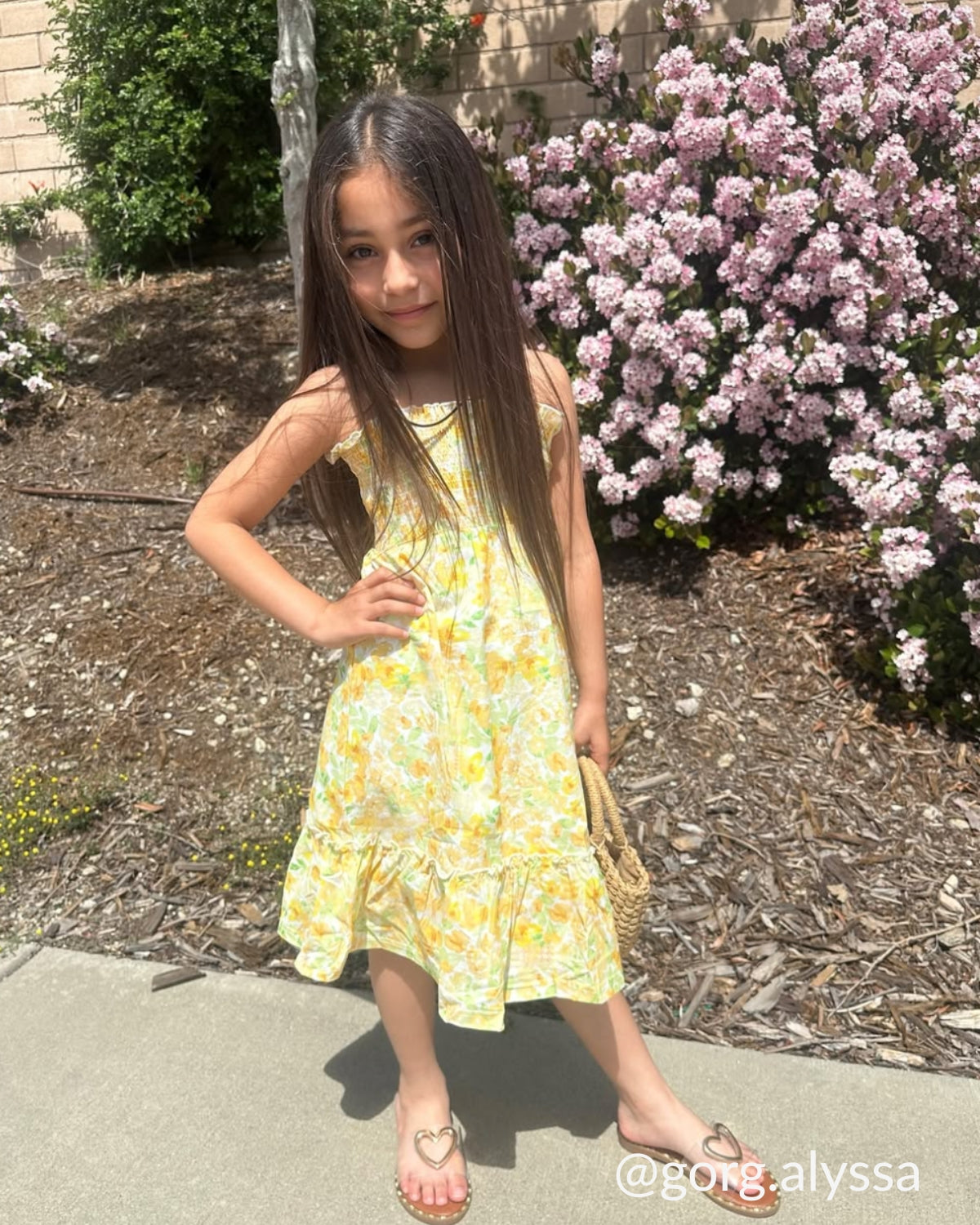 Young girl in a yellow floral dress standing outdoors with pink flowers in the background