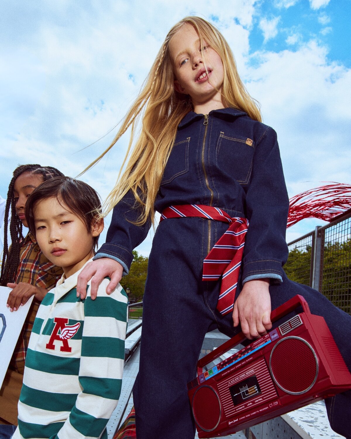 Three children outdoors with a clear blue sky, one holding a red boombox.