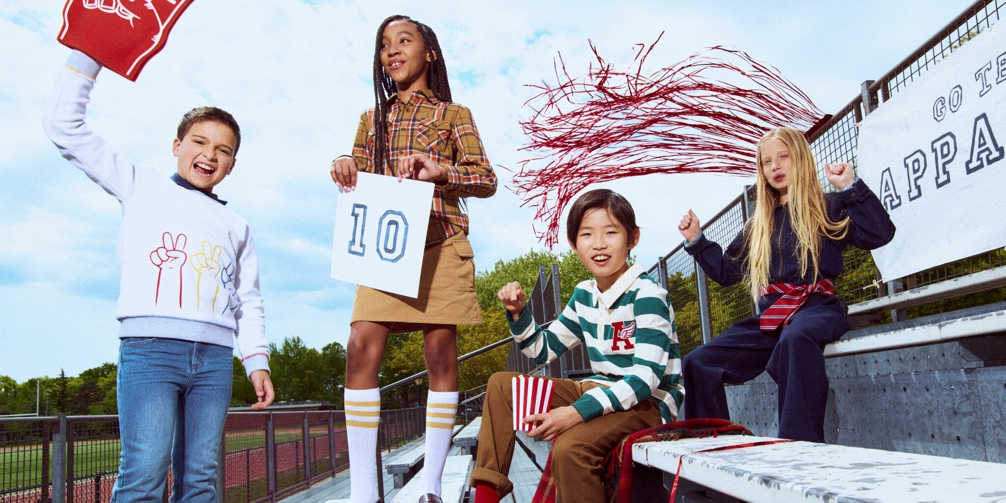 Four children in a park with one holding a sign with the number 10, another with a red pom-pom, and two others sitting on a bench.