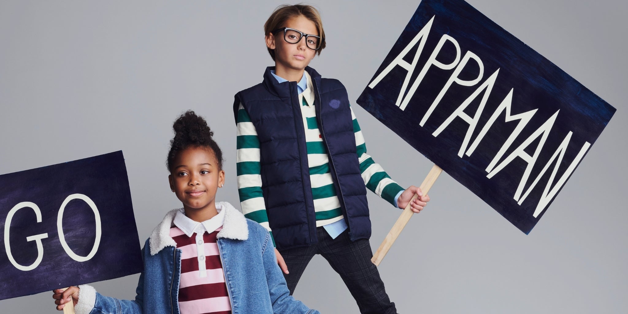 Two children modeling streetwear outfits and holding large navy signs with white text that read 'GO' and 'APPAMAN' against a light gray studio background.